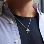 A close-up of a person wearing a silver pendant necklace with a cross and a small circular medallion, styled over a navy blue t-shirt and a light blue button-down shirt.