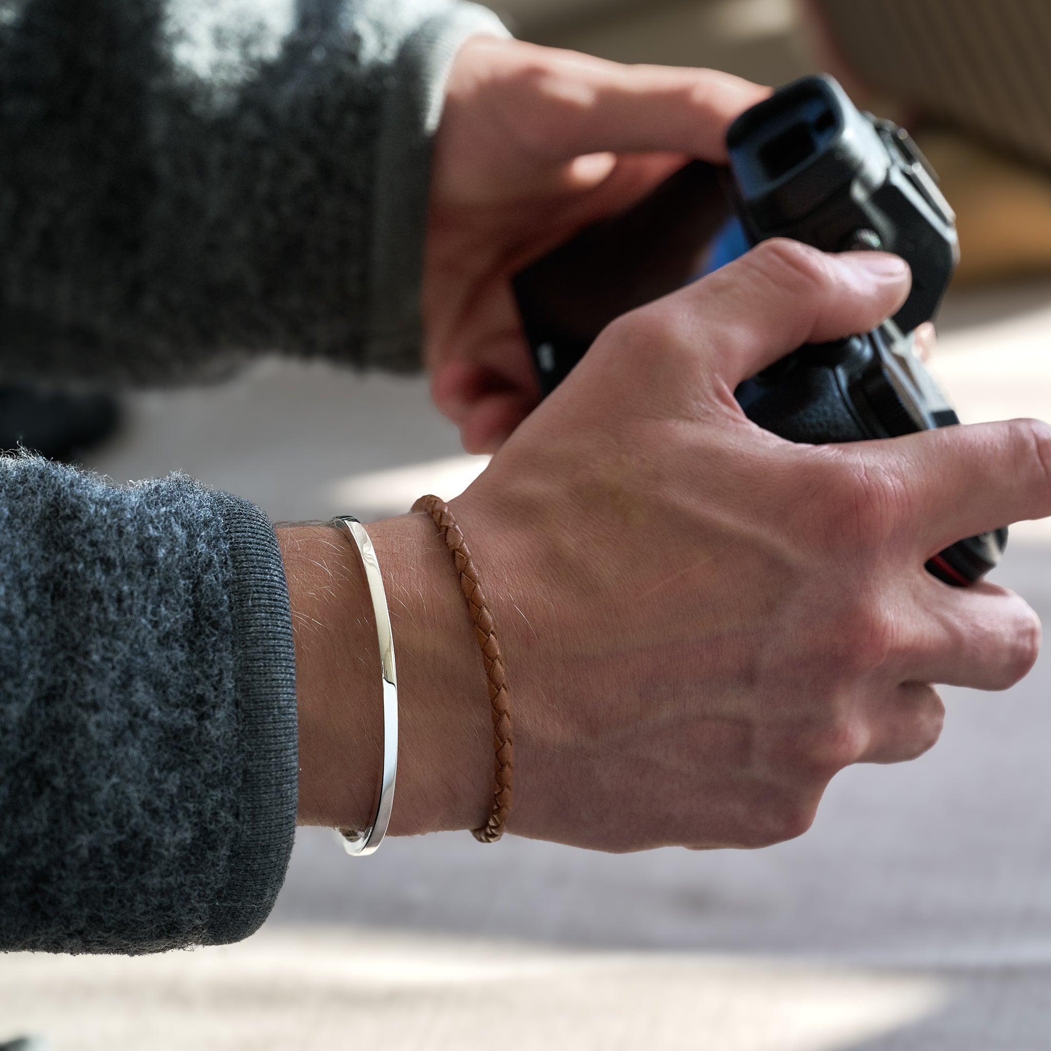 Close-up of hands holding a camera with silver bracelet and brown plaited bracelet on wrist and a blurred background