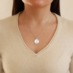 Silver necklace with a round pendant worn by a person in a beige sweater.