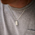 Person wearing a grey shirt with a necklace featuring a rectangular pendant.