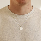 Person wearing a silver necklace with a round pendant on a plain background