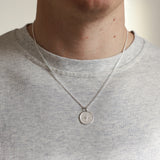 Person wearing a silver necklace with a round pendant on a plain background