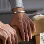 Close-up of a person's hands resting on a wooden chair wearing a silver hammered bracelet with a blurred background