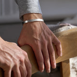 Close-up of a person's hands resting on a wooden chair wearing a silver hammered bracelet with a blurred background