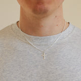 Person wearing a silver necklace with a cross pendant on a plain background