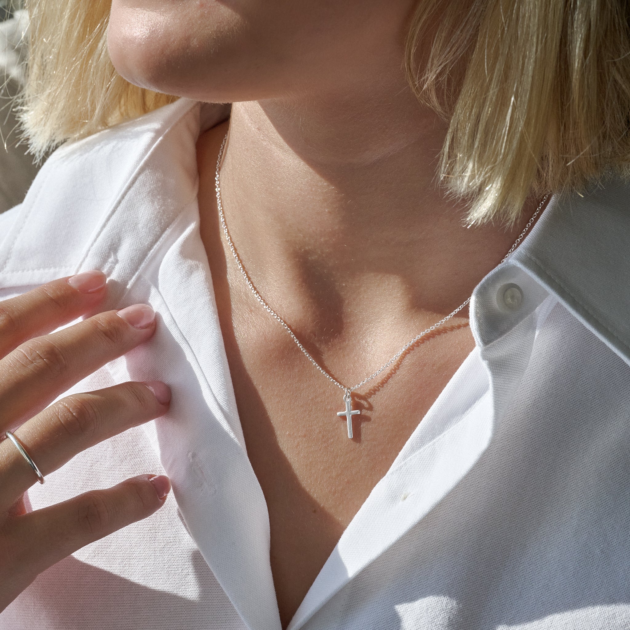 Close-up of a person in a white shirt wearing a silver necklace with a cross pendant, against a blurred background.