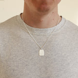 Person wearing a silver necklace with a rectangular pendant on a plain background
