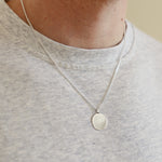Person wearing a silver necklace with a round pendant on a plain background