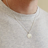 Person wearing a silver necklace with a round pendant on a plain background