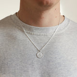 Person wearing a silver necklace with a round pendant on a plain background