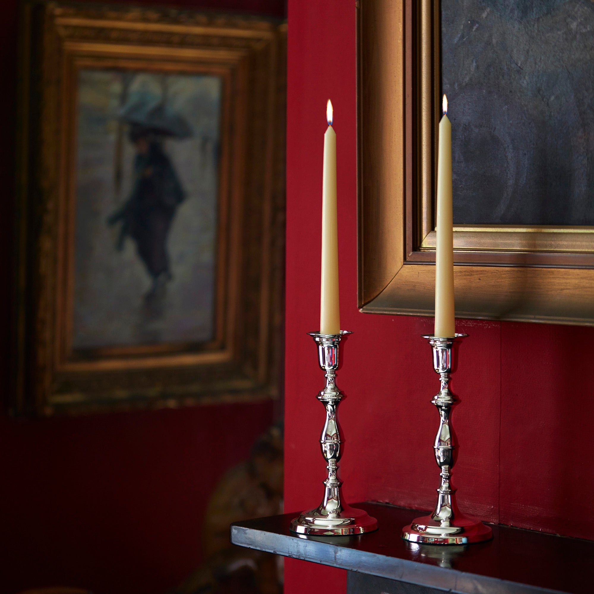 Two candlesticks with candles on a red wall with framed artwork.
