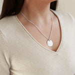 Silver necklace with a round pendant worn by a person in a beige top.